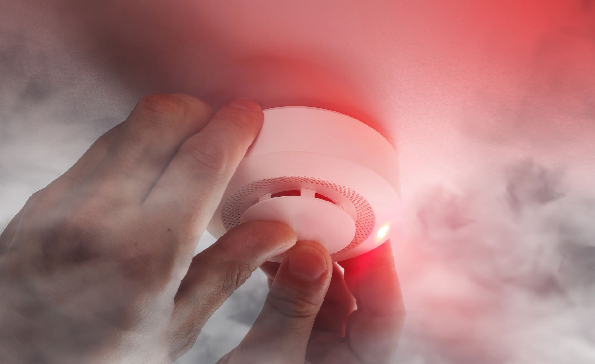 Hands reaching for a smoke detector, surrounded by smoke; red light flashing.