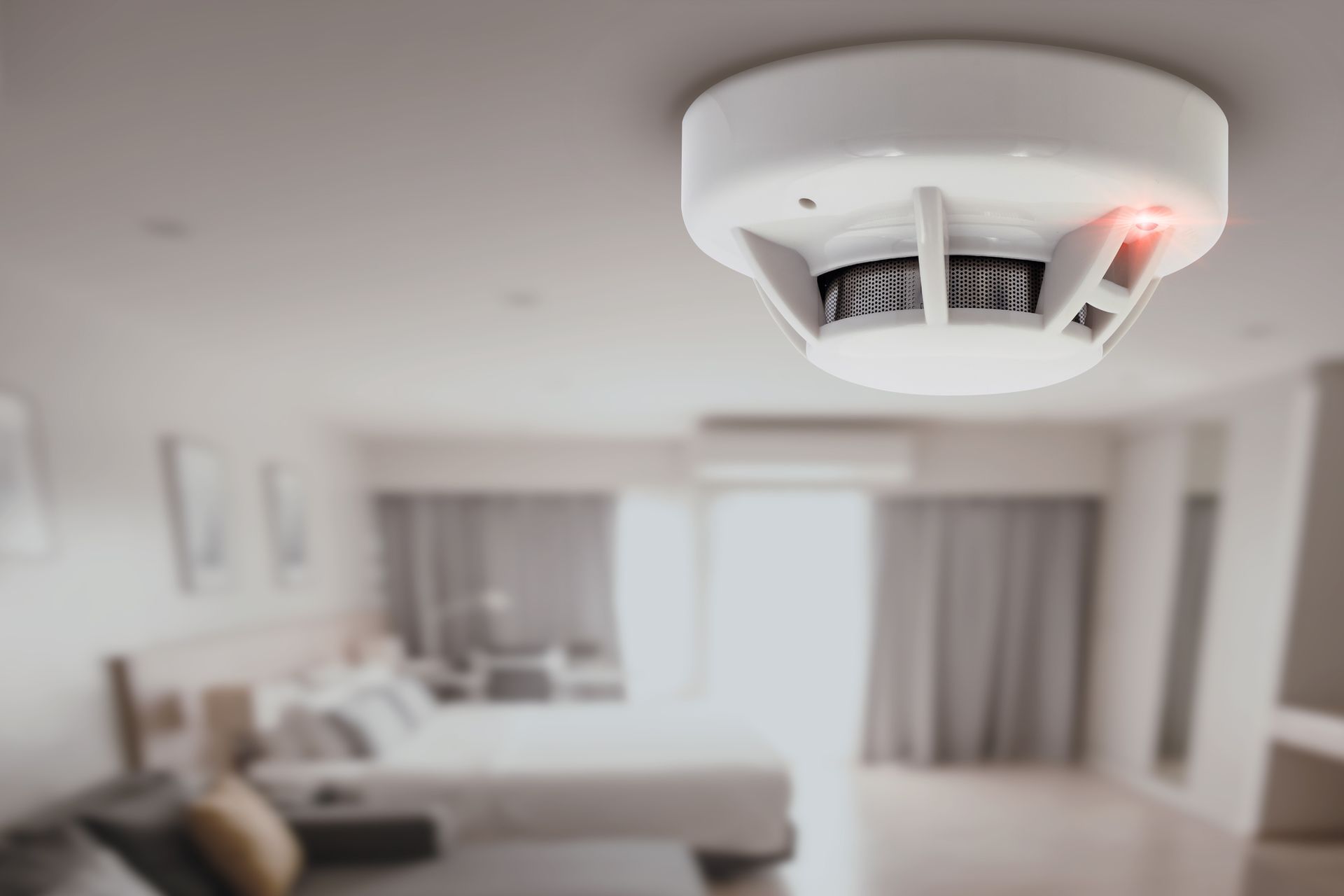 White smoke detector on a ceiling with a red light on, in a blurry bedroom. White smoke detector on a ceiling with a red light on, in a blurry bedroom.