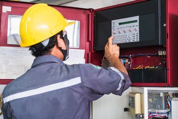 Hand reaching to activate a red fire alarm box on a white wall; 