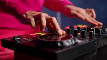 Hands adjusting knobs on a black DJ controller with a glowing jog wheel, person in a pink top nearby
