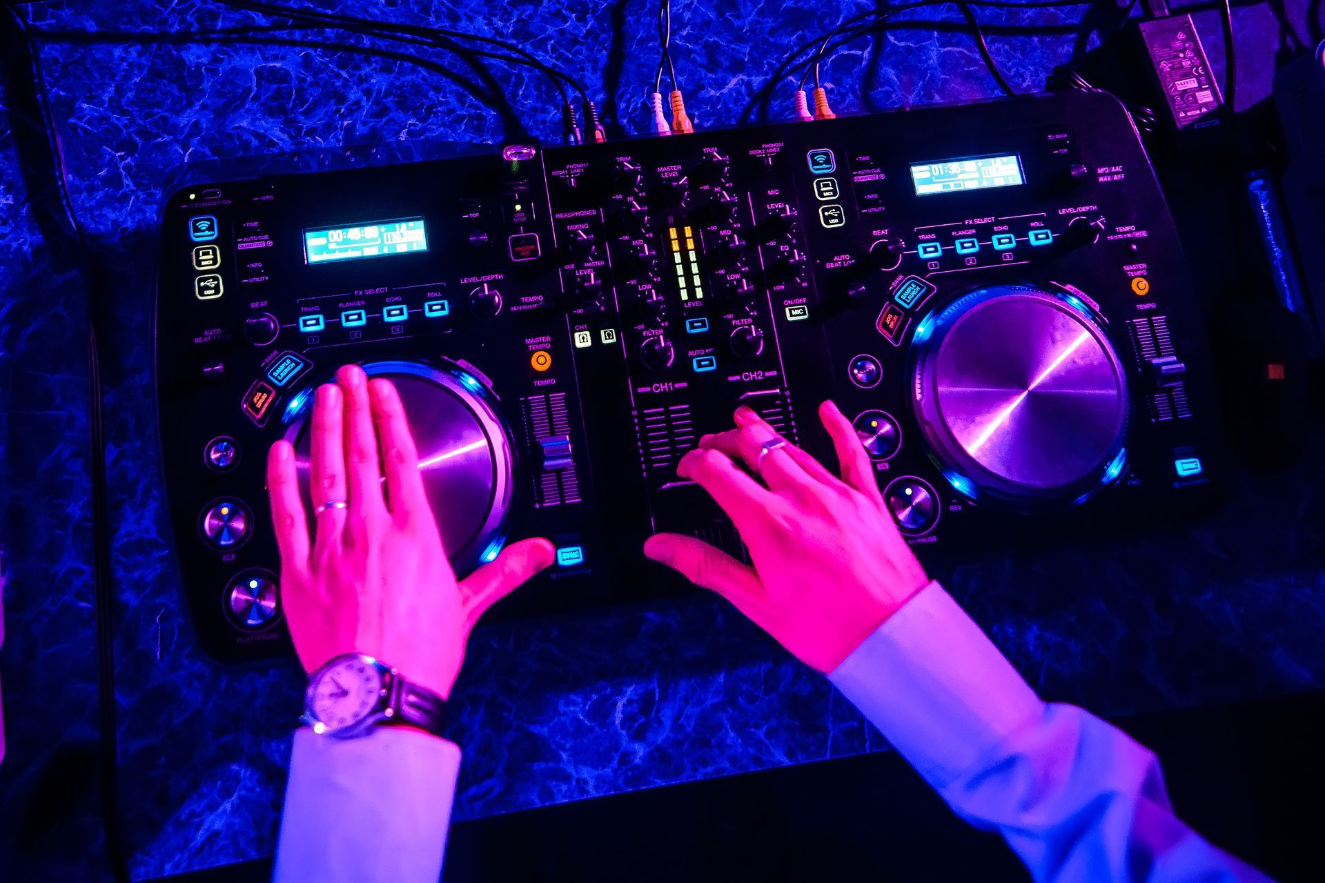 Hands adjust DJ turntables and mixer glowing blue and pink under neon lighting.