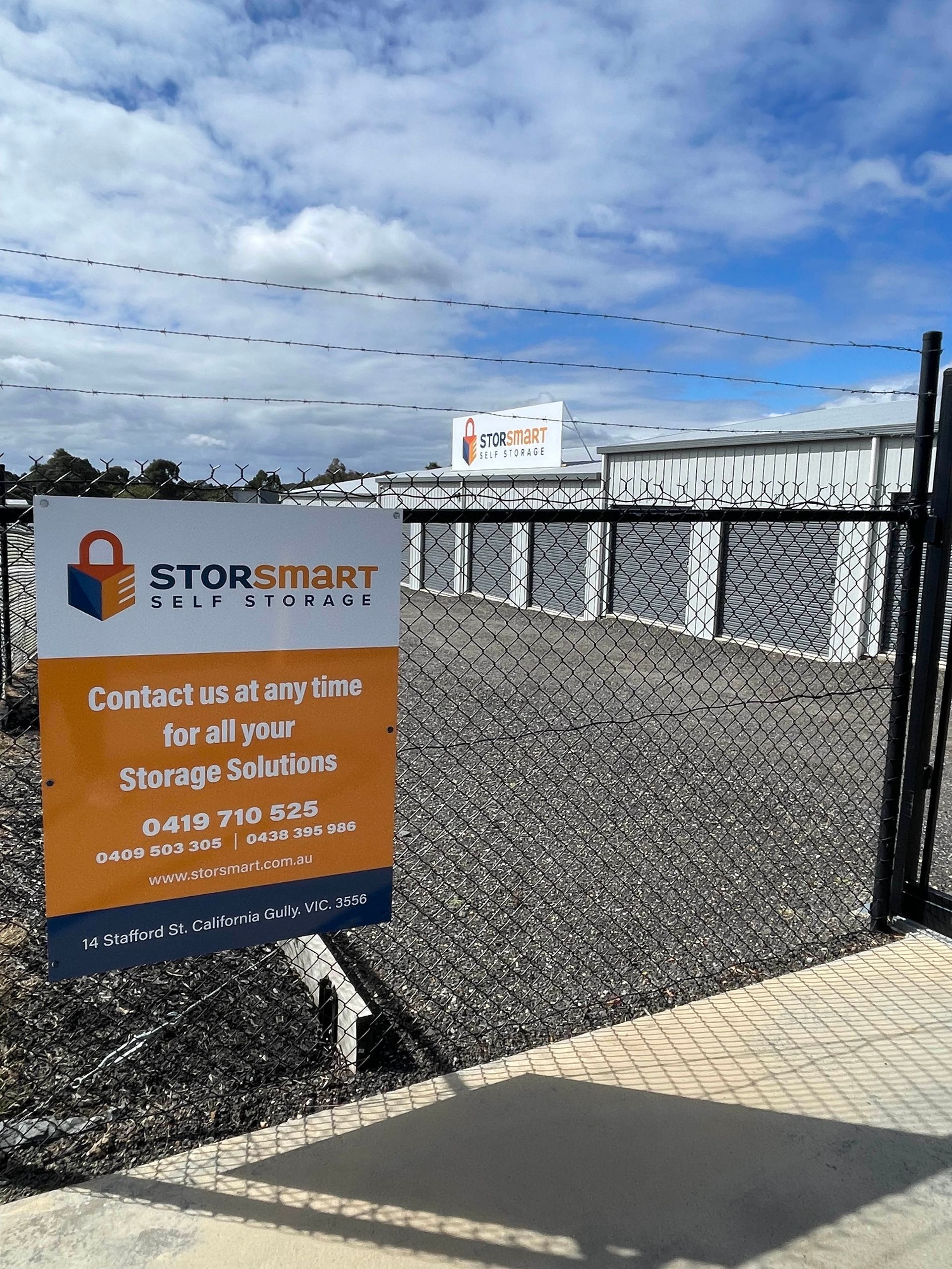 Storage facility entrance with Stop Smart sign, black chain-link fence, gravel lot, and blue sky