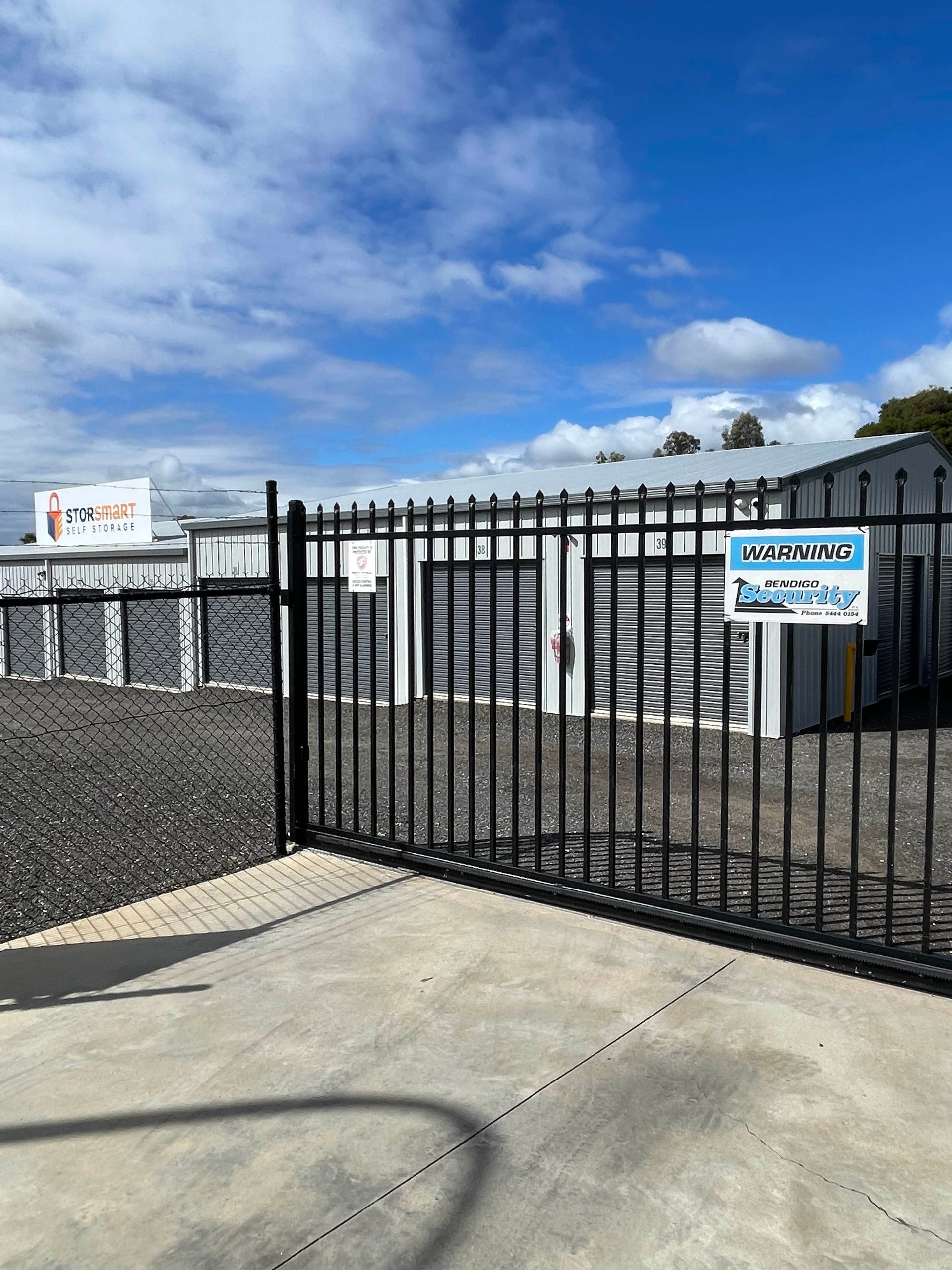Black metal gate and fence at a gravel lot under a blue sky, with a white building and sign in the background