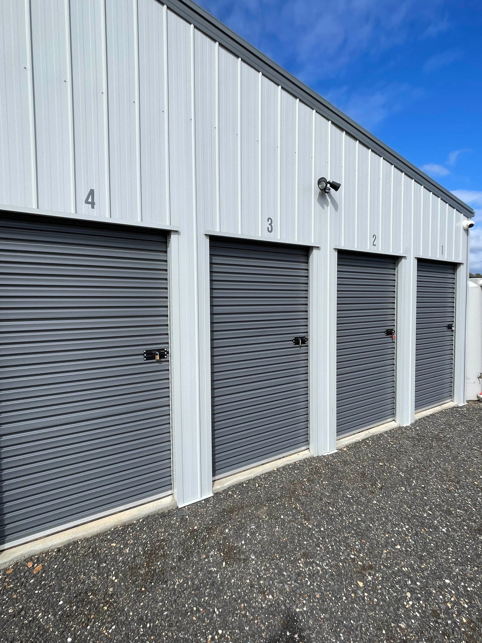 Row of gray storage unit doors in a metal building on a gravel lot under a blue sky