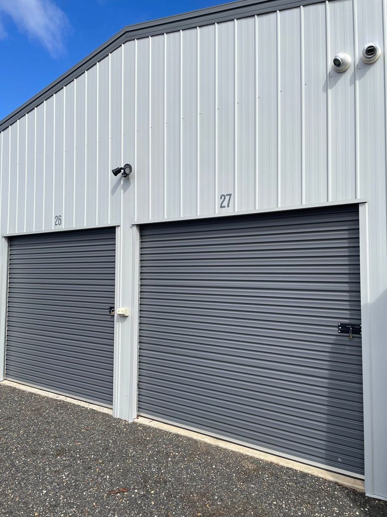 Gray industrial building with two closed roll-up garage doors and a gravel foreground