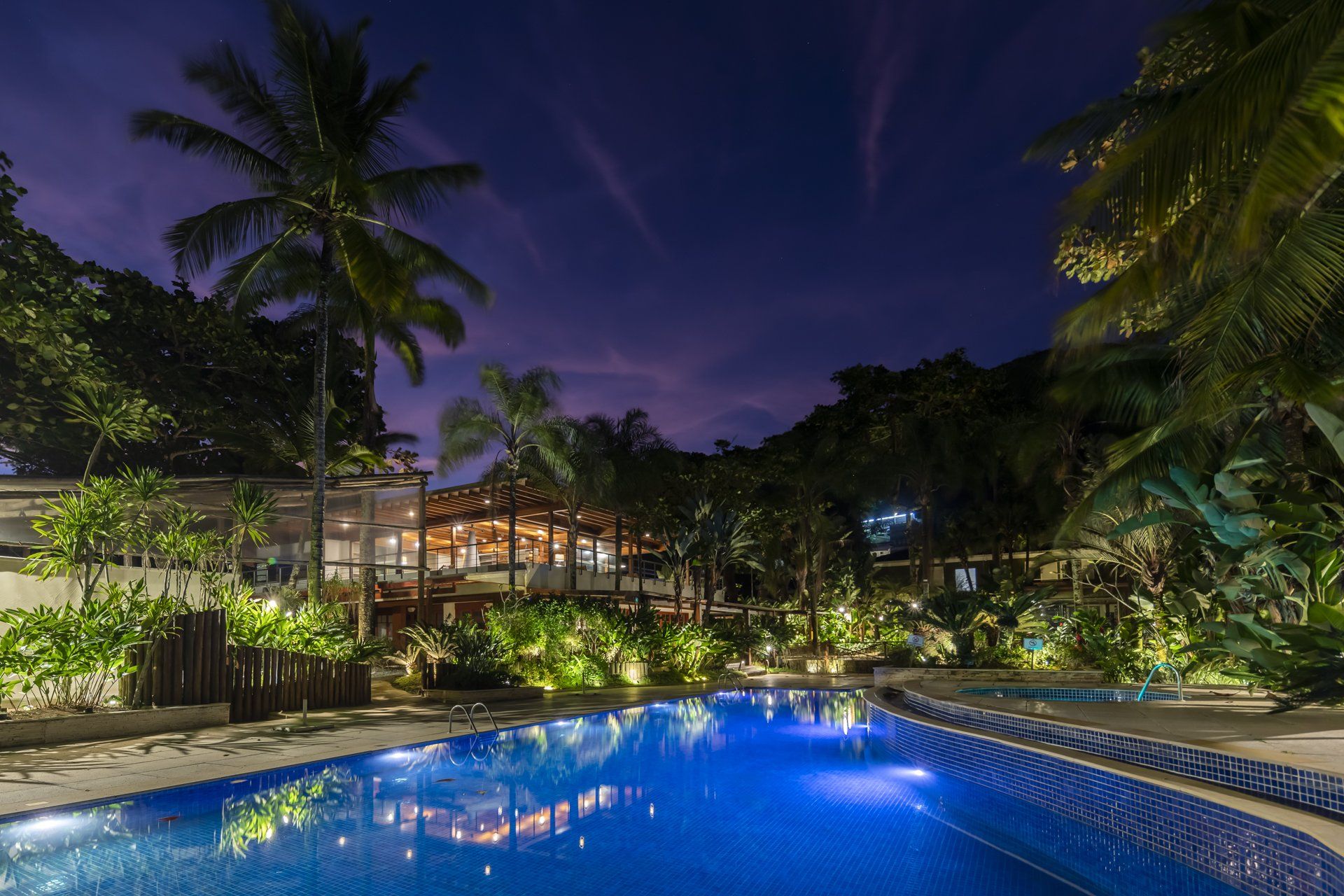 Uma grande piscina é iluminada à noite com palmeiras ao fundo.
