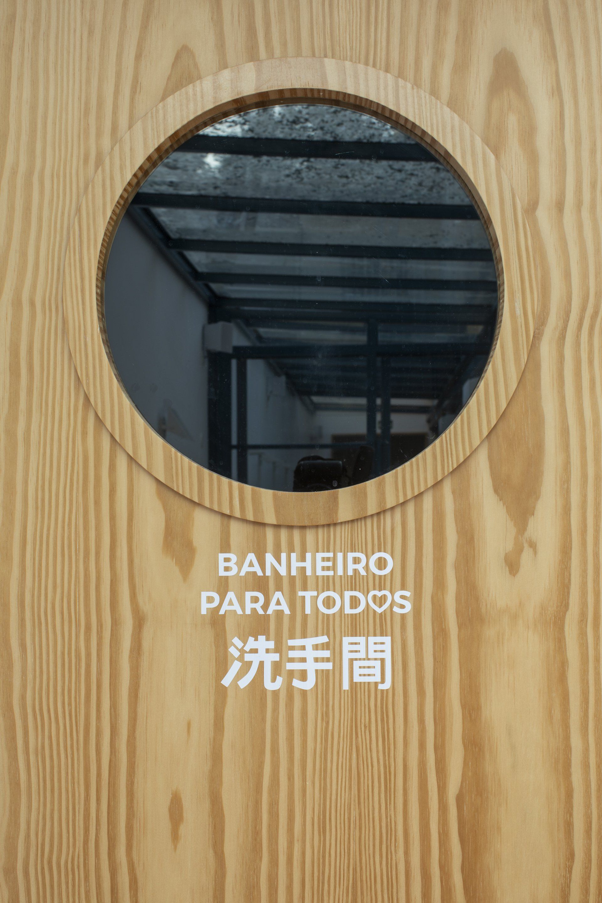 Uma porta de madeira com janela redonda e nela escritas as palavras banheiro para todos.
