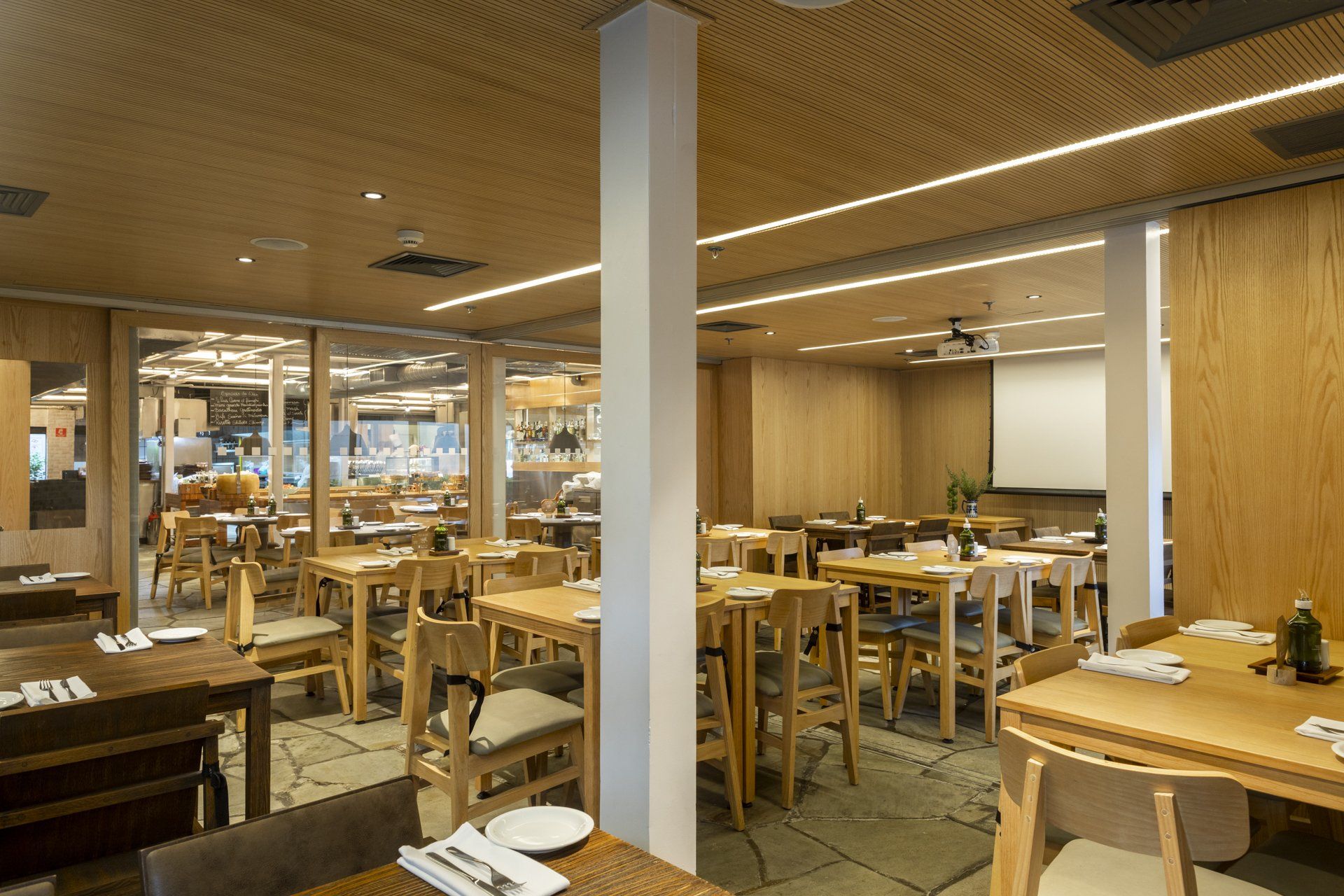 Um restaurante com mesas e cadeiras de madeira e tela de projeção.