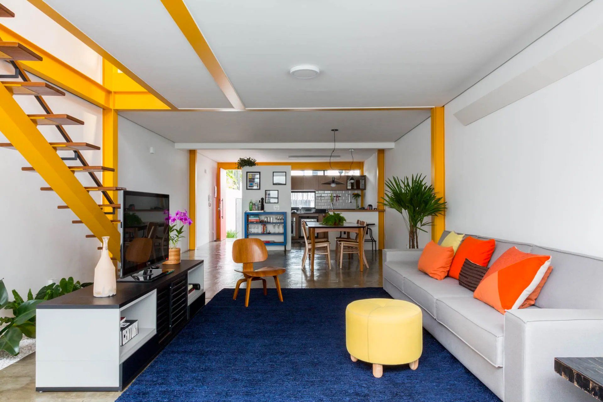 Uma sala com sofá, pufe, televisão e escadas.