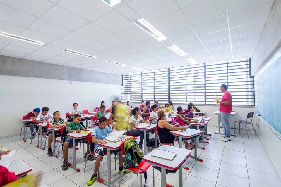 Um grupo de crianças está sentado em carteiras em uma sala de aula com um professor.