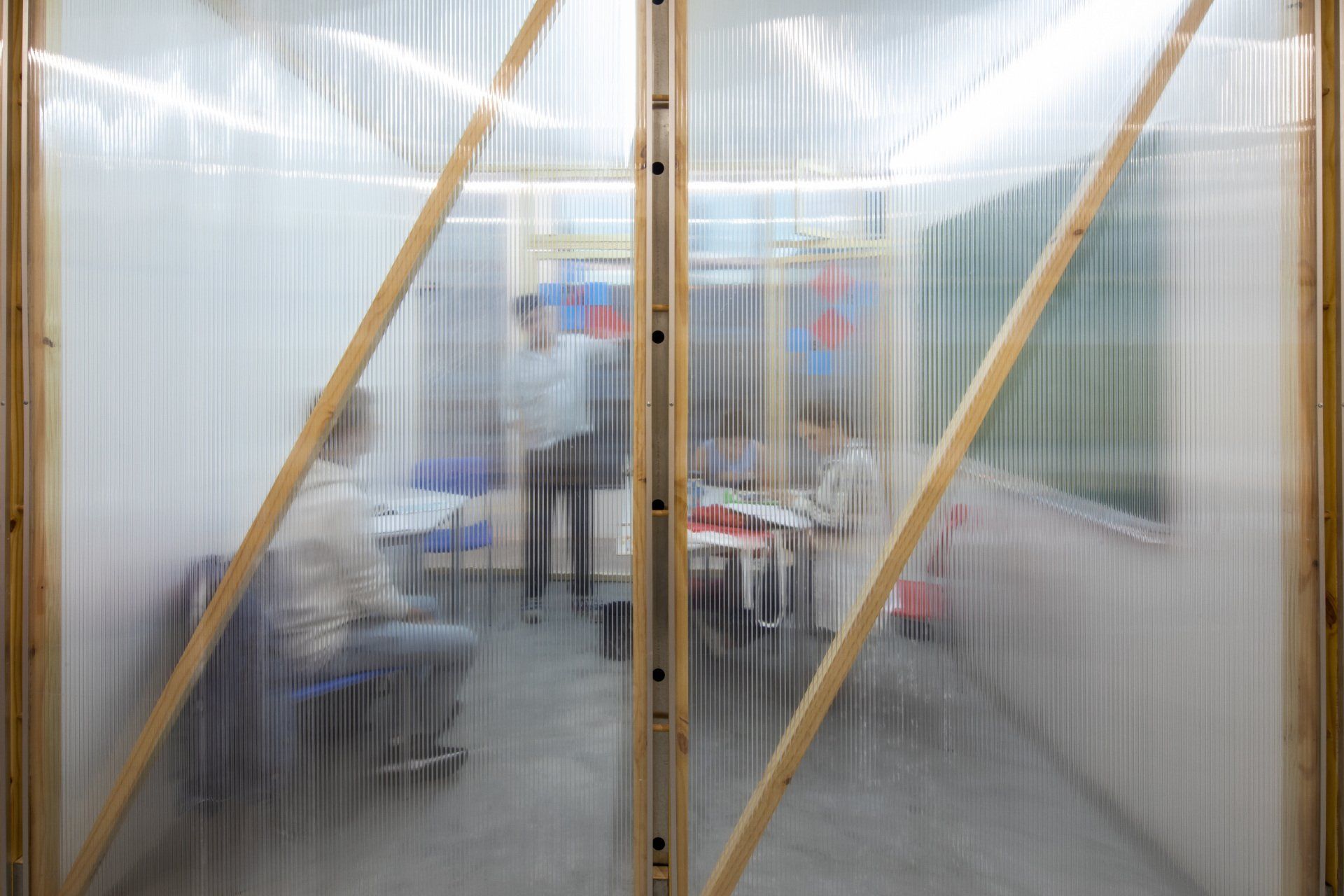 Um grupo de pessoas está sentado em mesas em uma sala atrás de uma parede de plástico transparente.