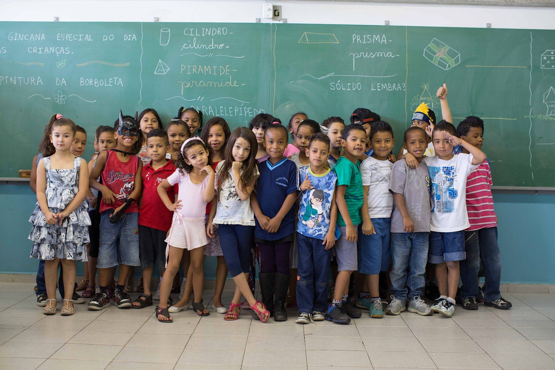Um grupo de crianças posa para uma foto em frente a um quadro negro em uma sala de aula.