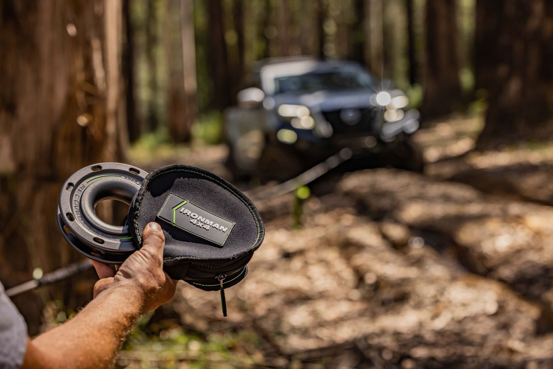 A person is holding a winch in front of a car in the woods.
