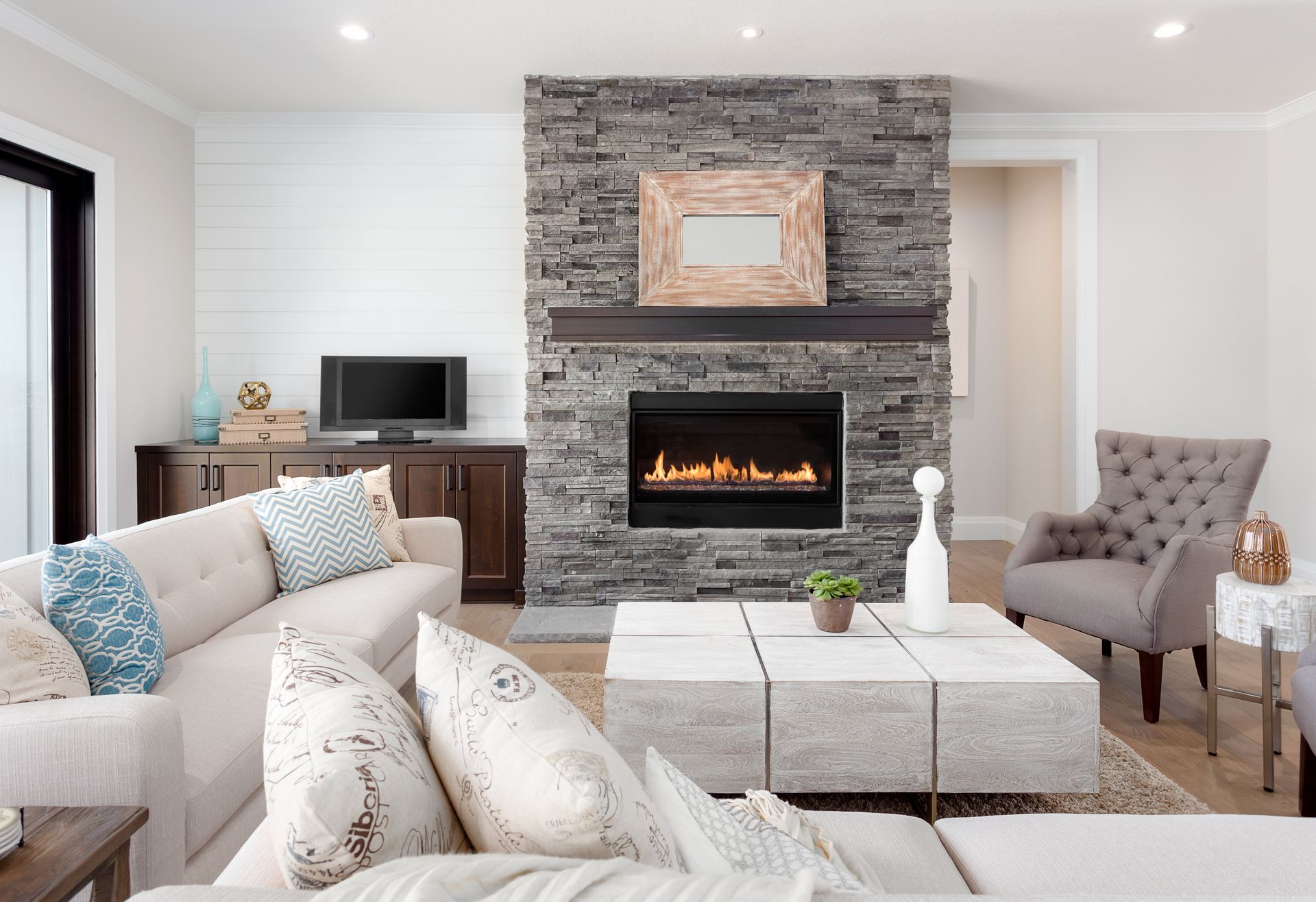 Cozy living room with gray stone fireplace, beige sofa, white ottomans, and dark wood desk