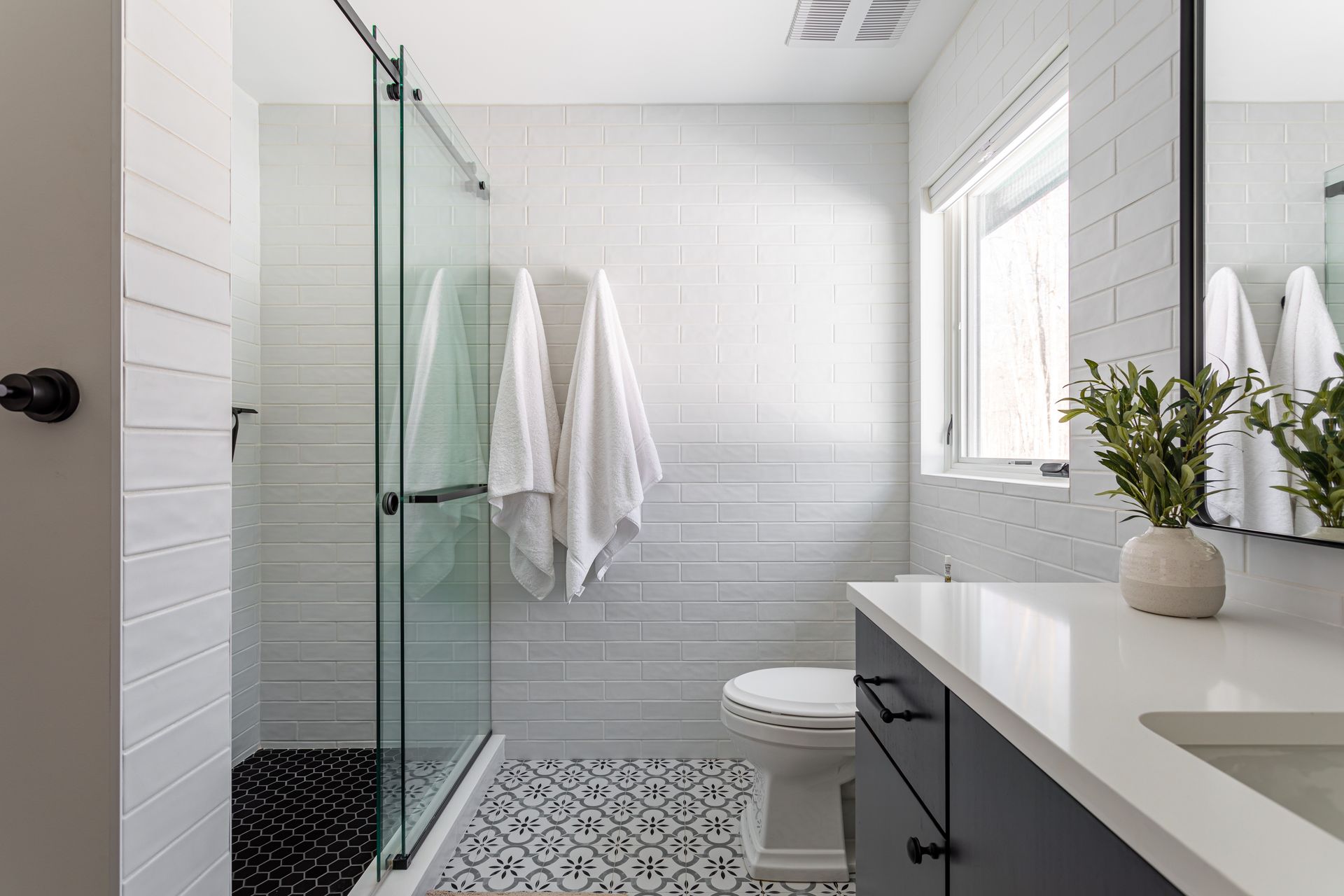 Modern white bathroom with glass shower, patterned floor tile, window, sink, and towels hanging on the wall Modern white bathroom with glass shower, patterned floor tile, window, sink, and towels hanging on the wall