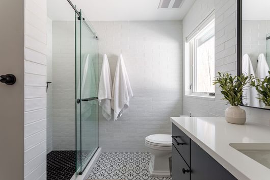Modern white bathroom with glass shower, patterned floor tile, window, sink, and towels hanging on the wall