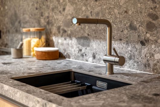 Modern kitchen sink with bronze faucet in a stone countertop against a textured stone wall