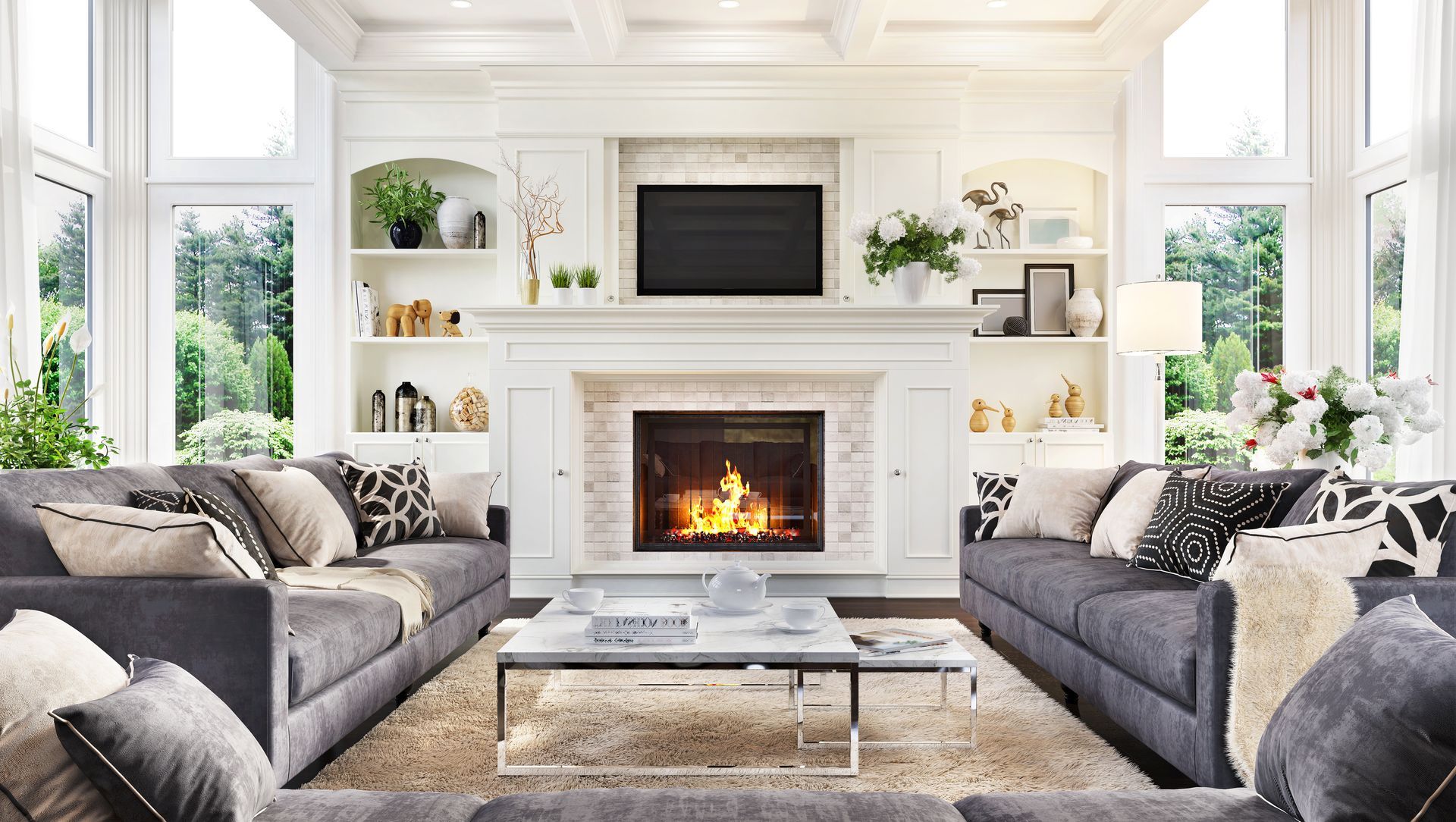 Bright living room with two gray sofas, a lit fireplace, white built-ins, and large windows.