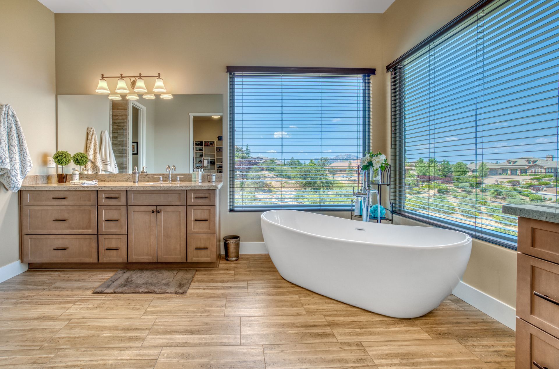 Bright bathroom with double vanity, freestanding tub, large windows, and wood-look floor.