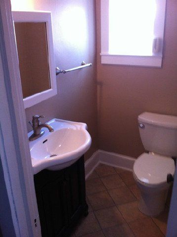 Small beige bathroom with sink, mirror, toilet, window, and tiled floor.