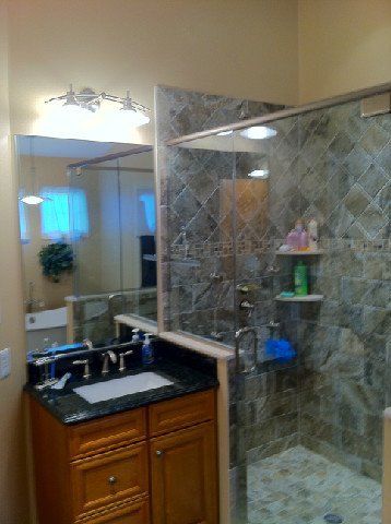 Bathroom vanity with granite counter, mirror, and glass shower stall with stone-pattern tile.