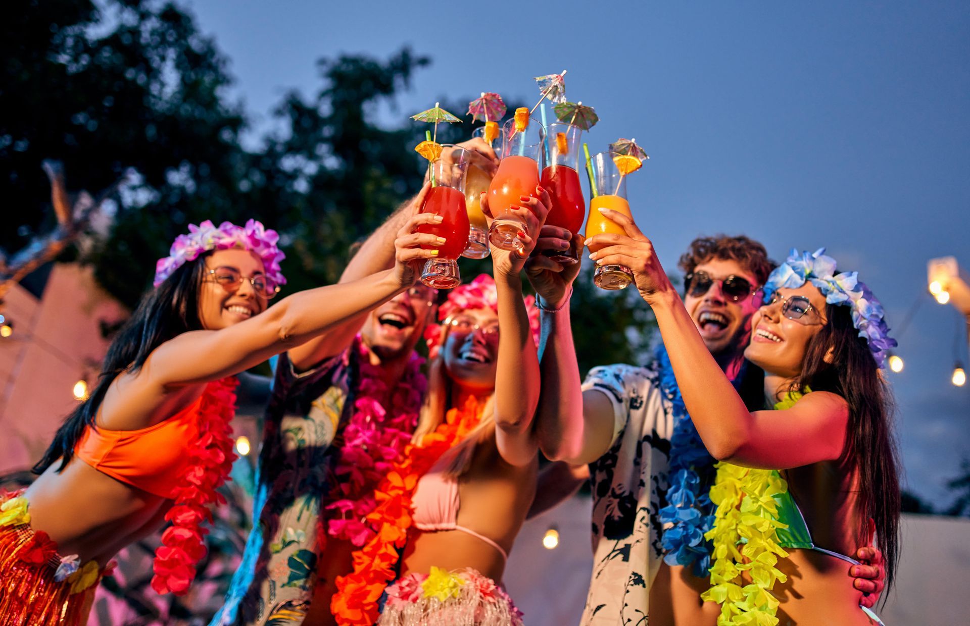 A group of people dressed in hawaiian costumes are holding up their drinks.