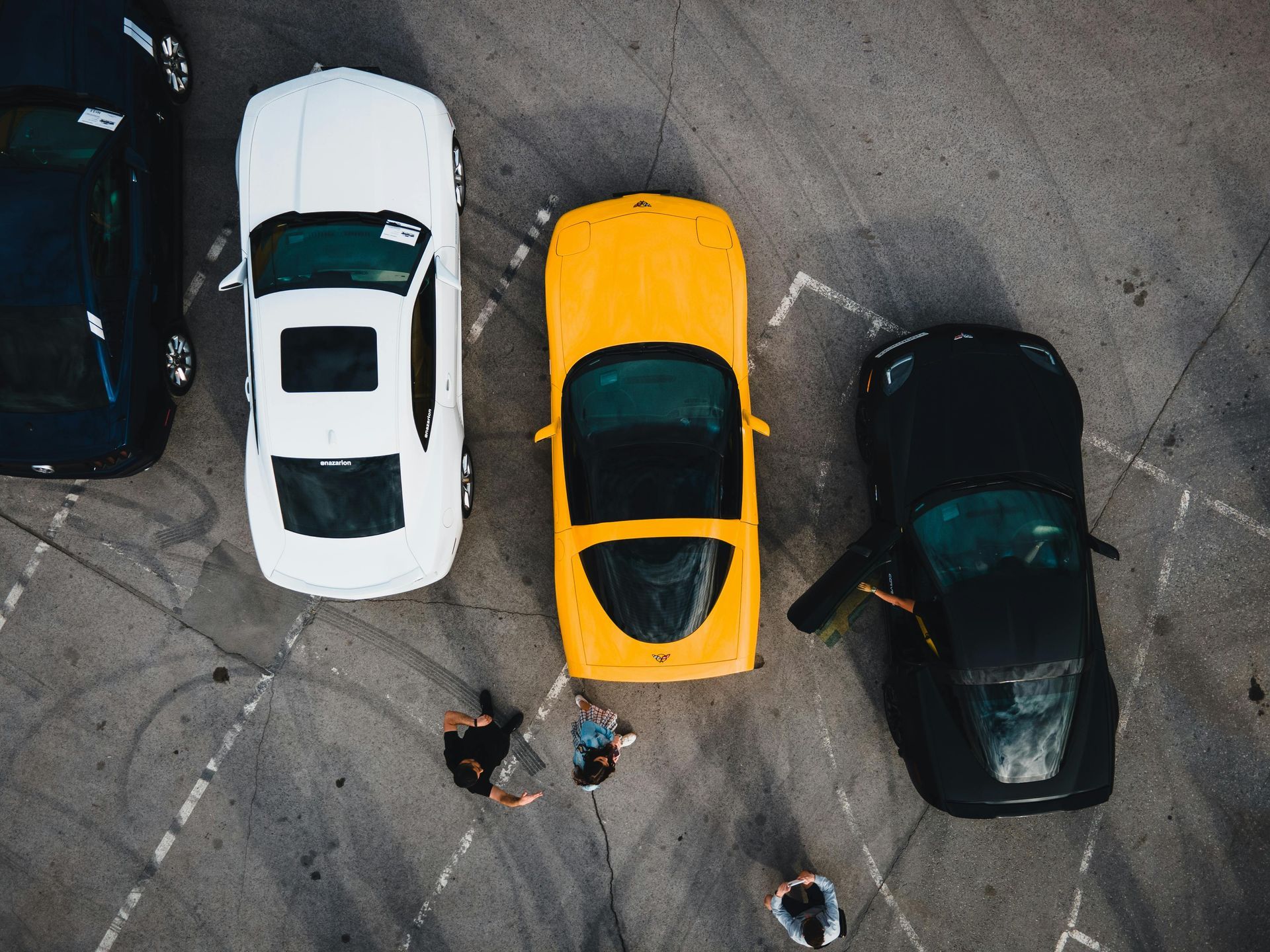 Overhead view of four cars parked on asphalt; two people standing near the yellow car.