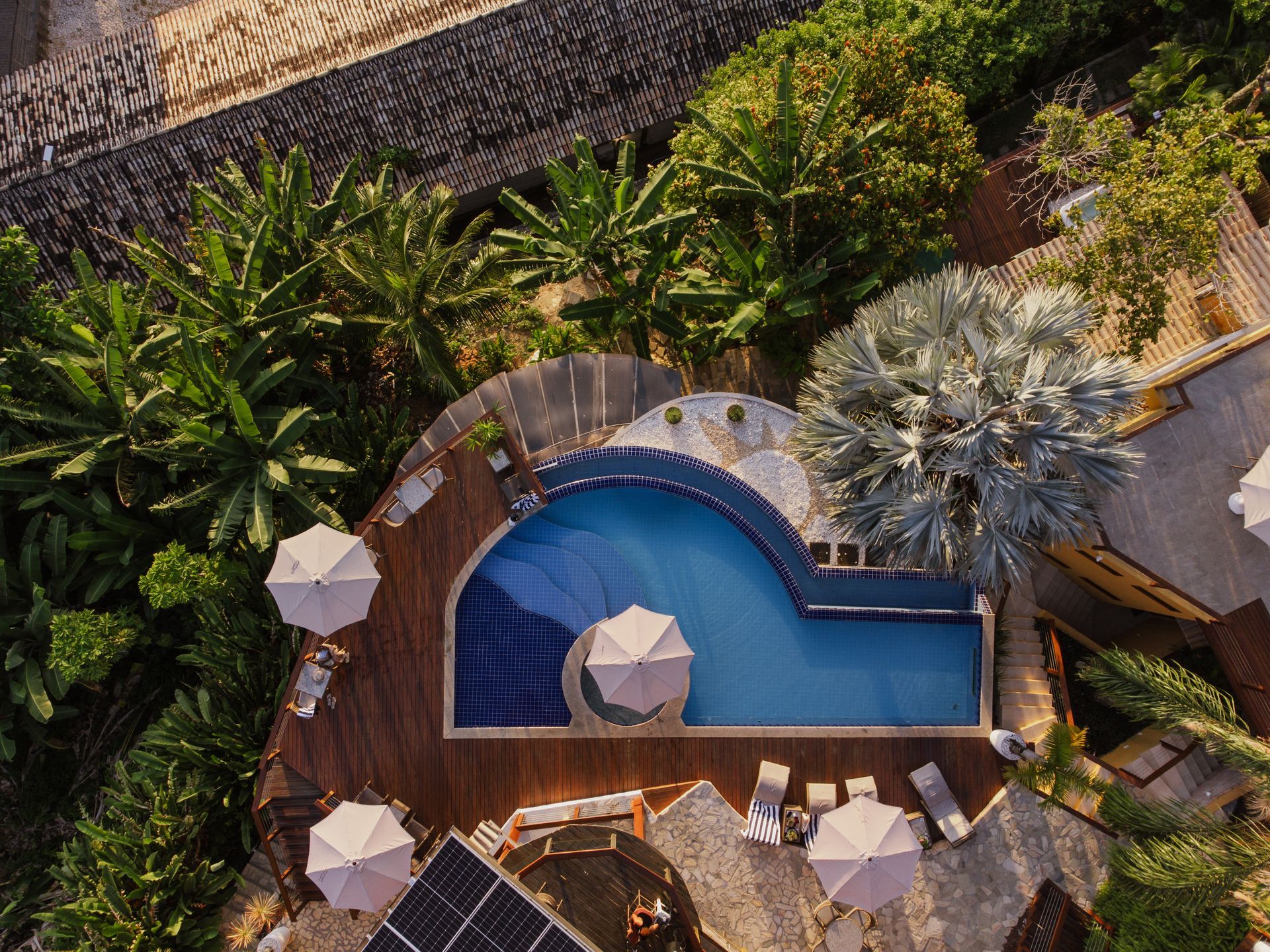 Vista aérea de uma piscina de resort cercada por árvores e espreguiçadeiras com guarda-sóis.