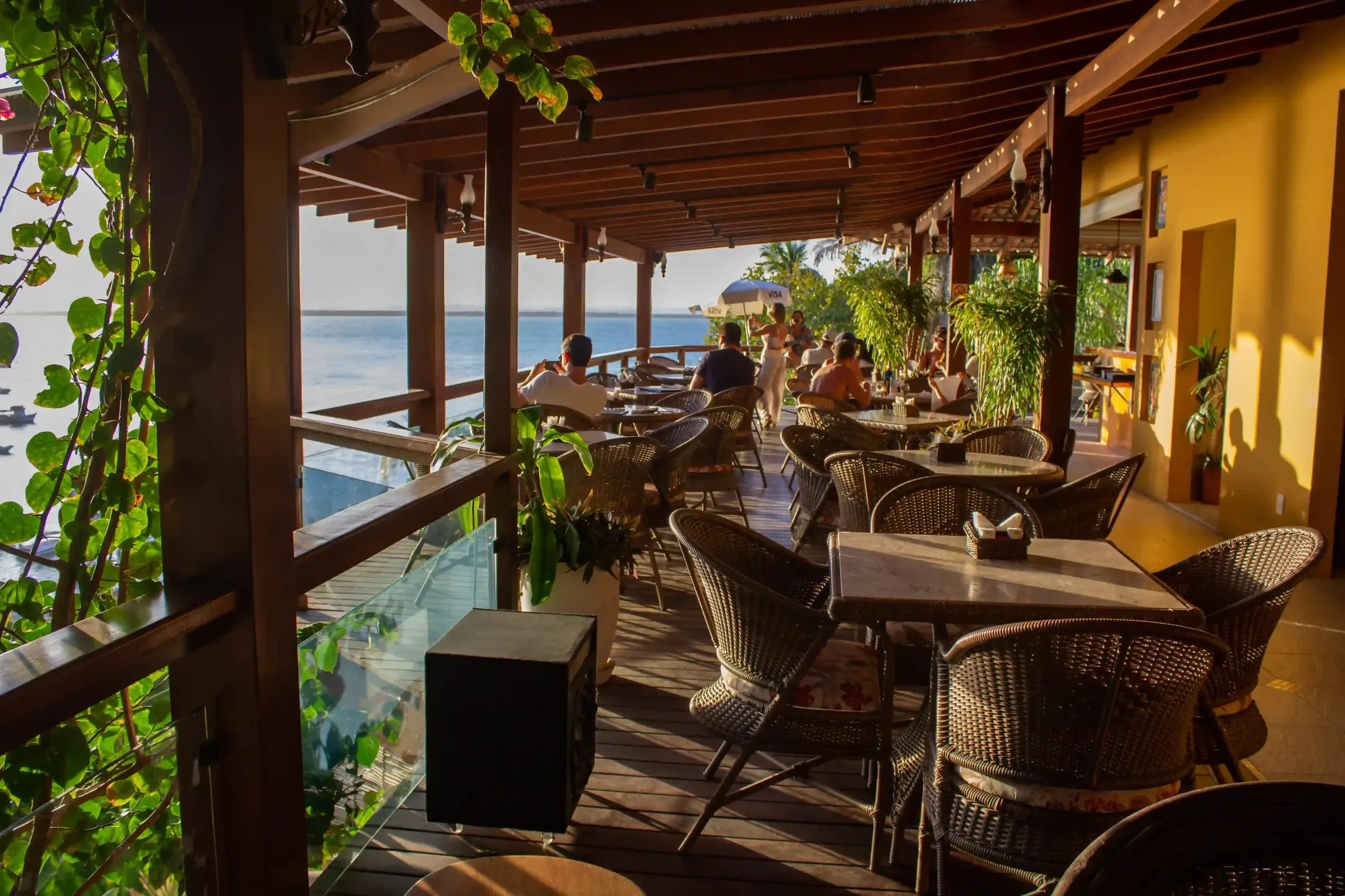 Pátio do restaurante com vista para a água, com mesas, cadeiras e clientes. Estrutura de madeira marrom, plantas verdes e paredes amarelas.