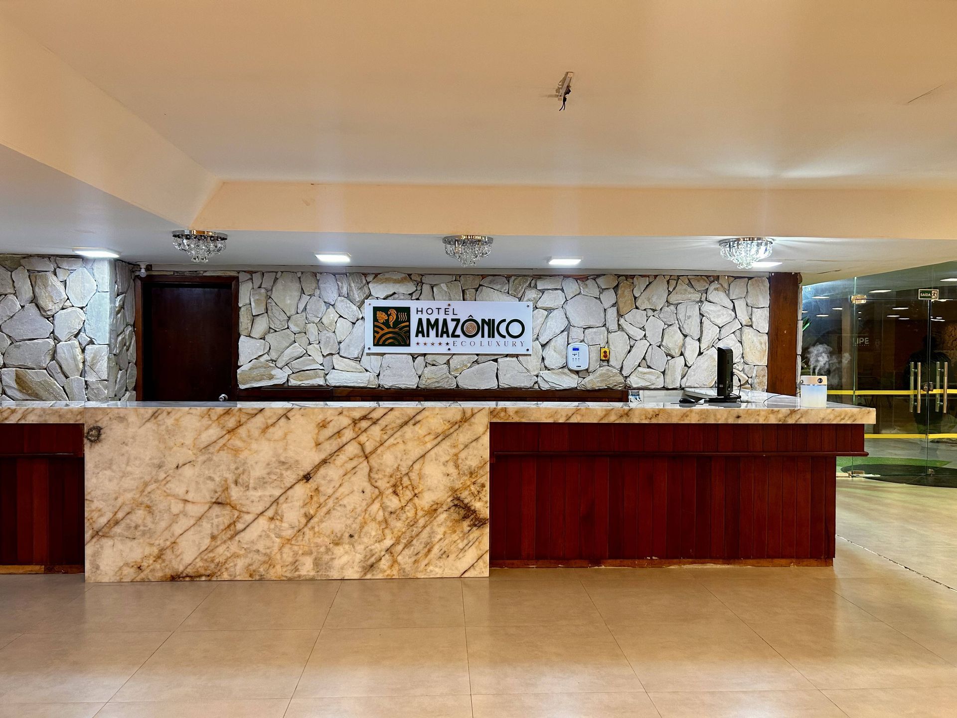 Balcão de recepção do hotel com detalhes em mármore e madeira, paredes bege e placa "Hotel Amazônico".