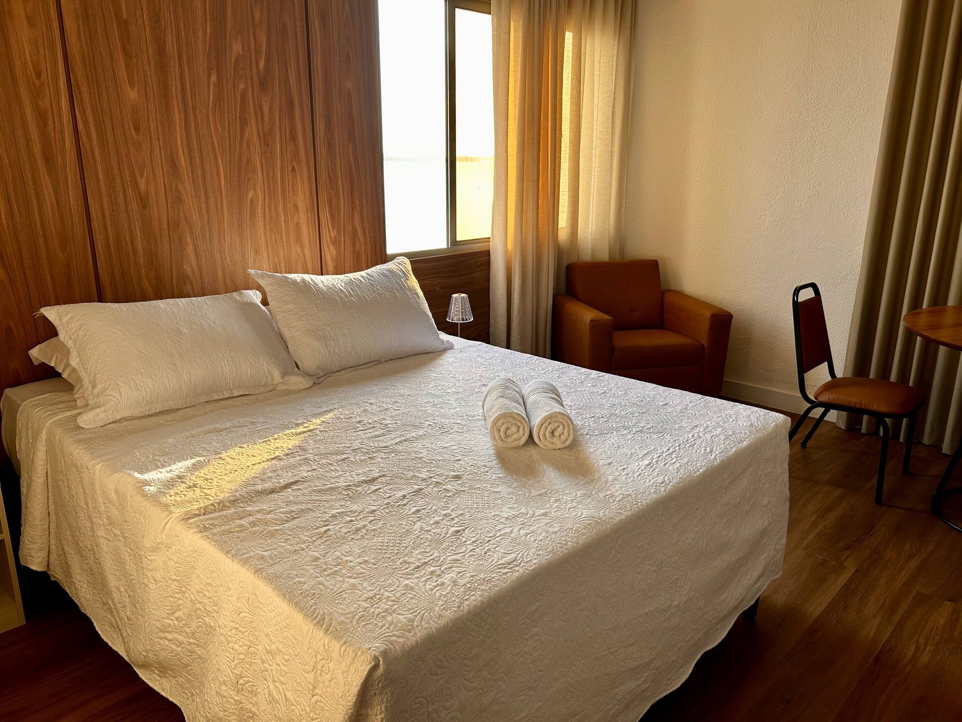 Quarto com cama, toalhas, cadeira e poltrona, próximo a uma janela com cortinas. Parede de madeira.