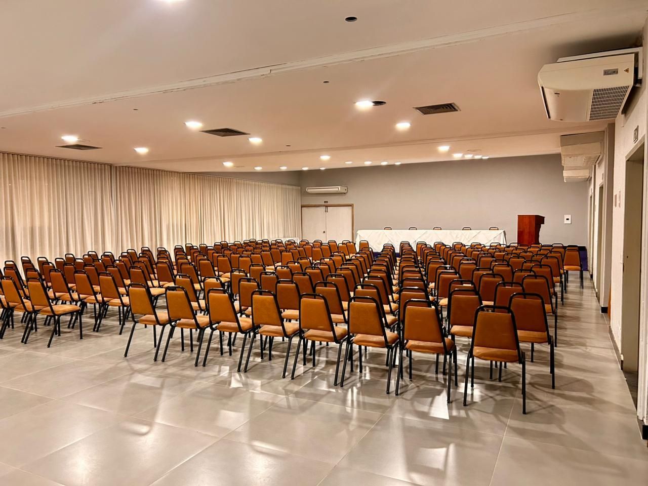 Sala de eventos vazia com fileiras de cadeiras voltadas para um palco com pódio.