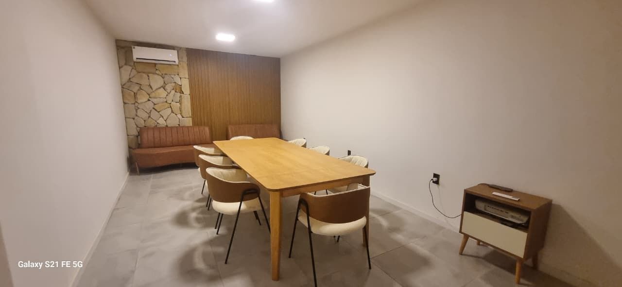 Sala de reuniões com uma longa mesa e cadeiras de madeira, um sofá, parede de pedra e um armário.