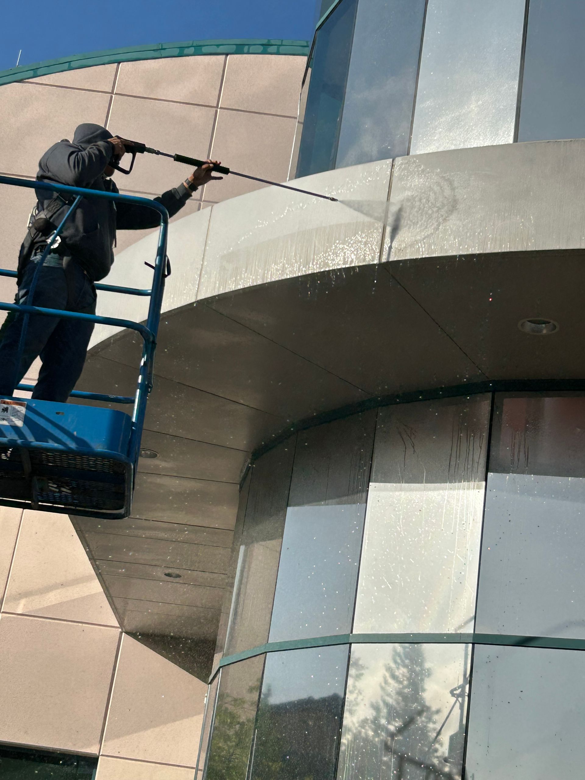 A person on a lift pressure washes a modern building's exterior. Blue sky background.