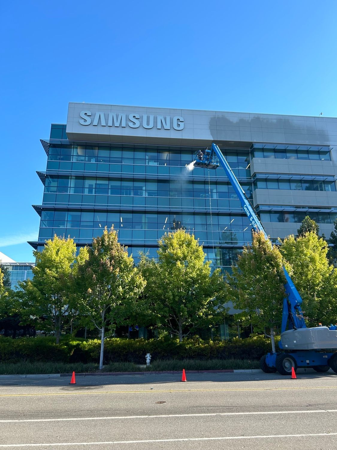 A worker on a lift is power washing a Samsung building’s facade. Blue lift, clear sky, green trees.