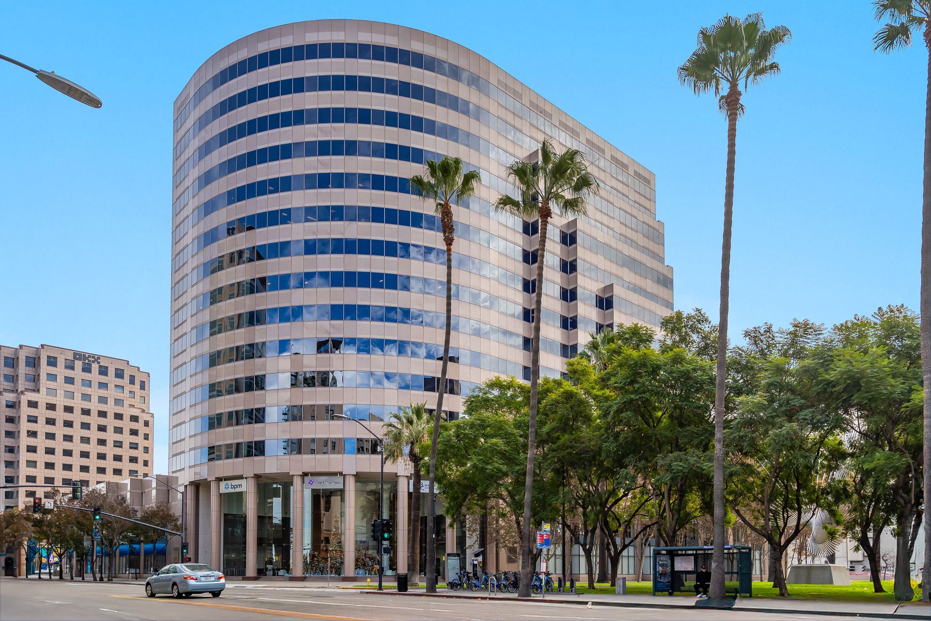 A tall, curved beige office building with glass windows, palm trees, and a street in front.