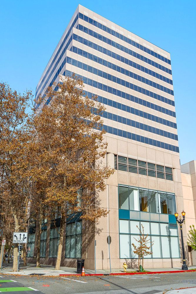 Tall modern office building with rows of windows and a beige facade, with trees and a sidewalk in front.