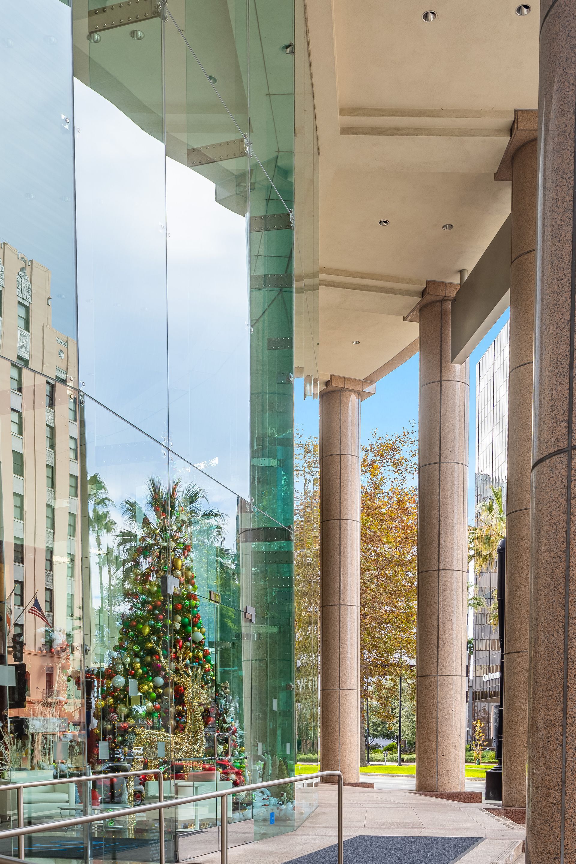 Exterior view with glass facade, columns, a Christmas tree reflected in the glass, and a blue sky.