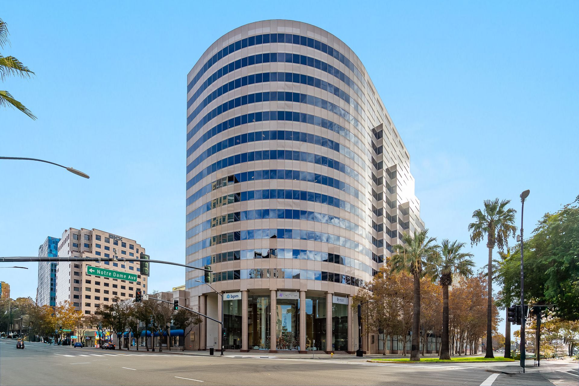 Curved, modern office building with many windows under a blue sky, palm trees nearby.