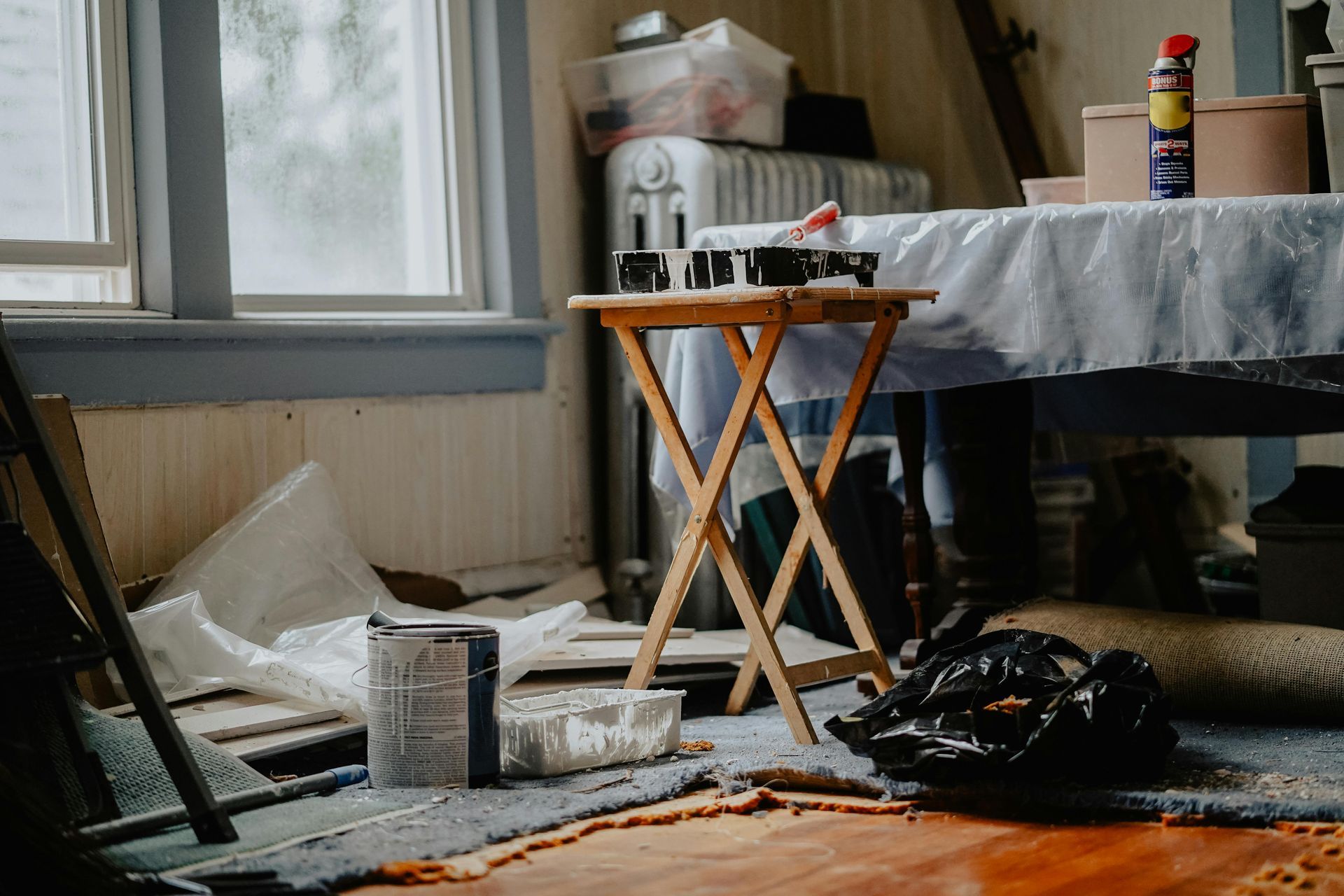 Messy room with paint supplies on a folding table, plastic-covered table, and trash bag on the floor near a window.