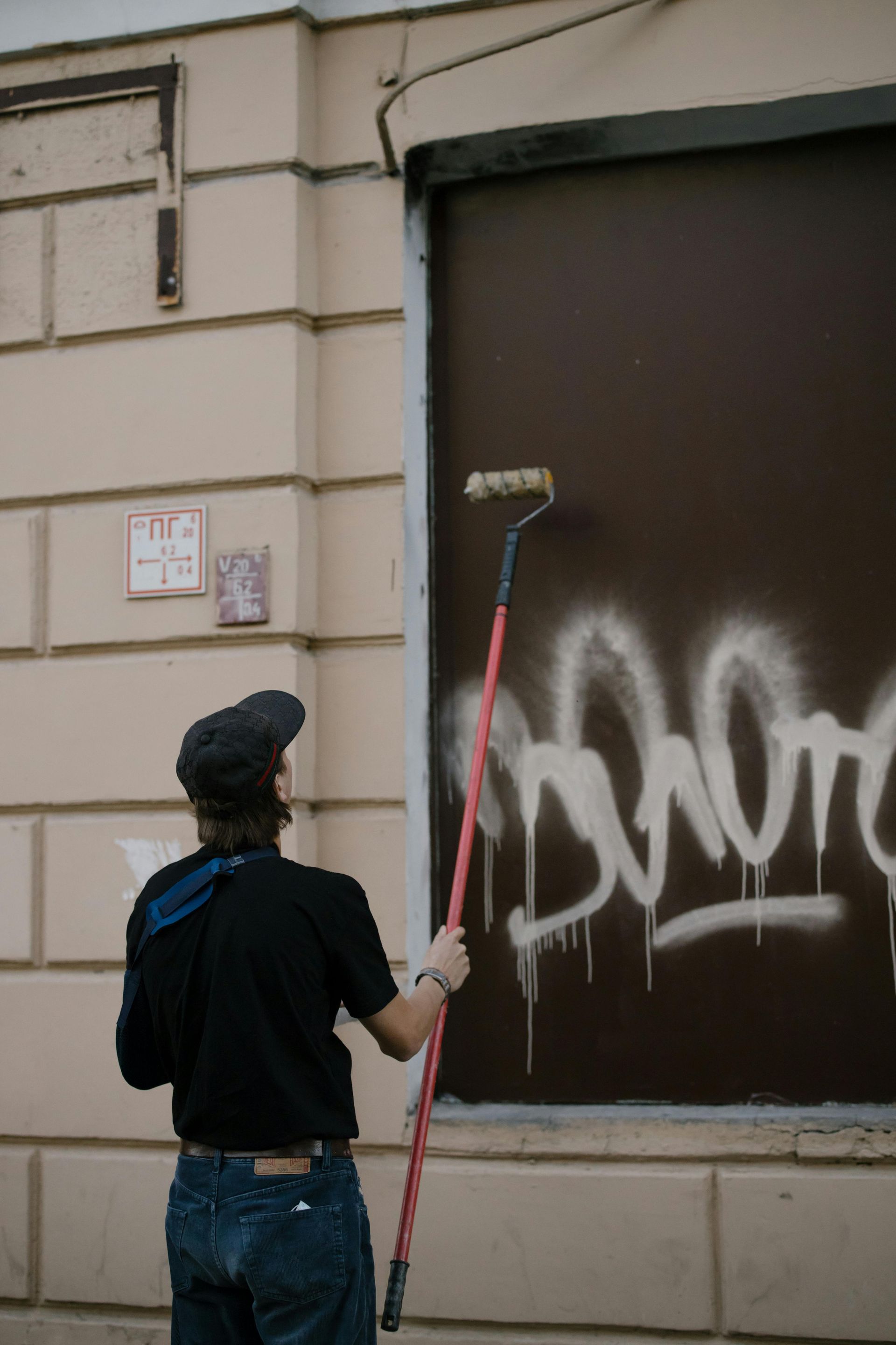 Person painting over graffiti on a dark surface with a roller, standing outside a building.