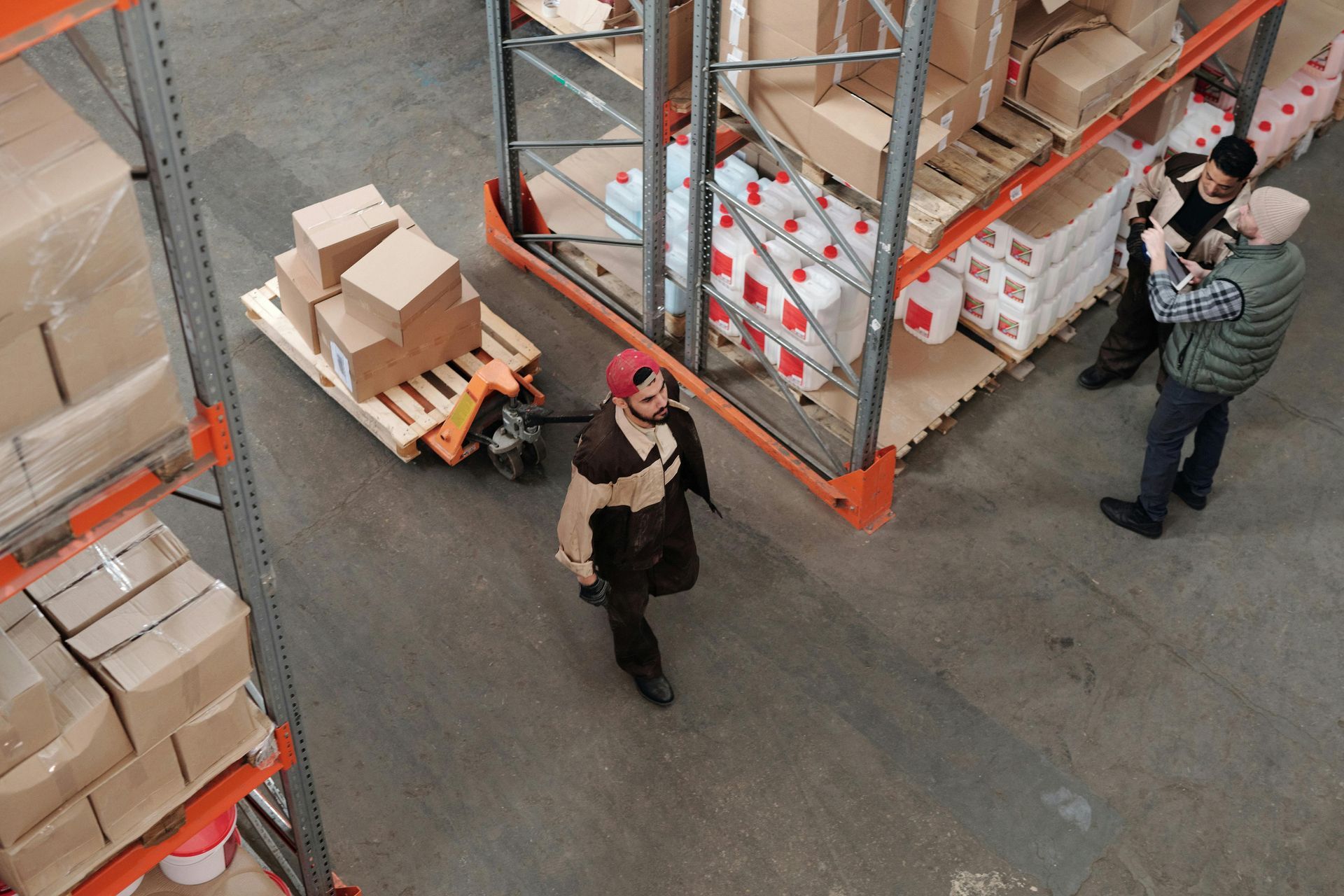 Warehouse workers near shelves of boxes. One worker pulls a pallet jack with boxes, others stand nearby.