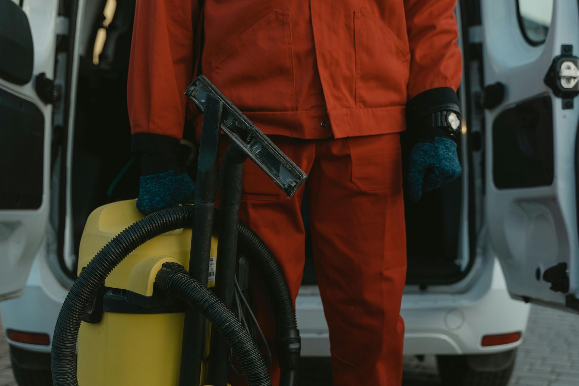 Person in red jumpsuit holding a vacuum and squeegee near an open vehicle.