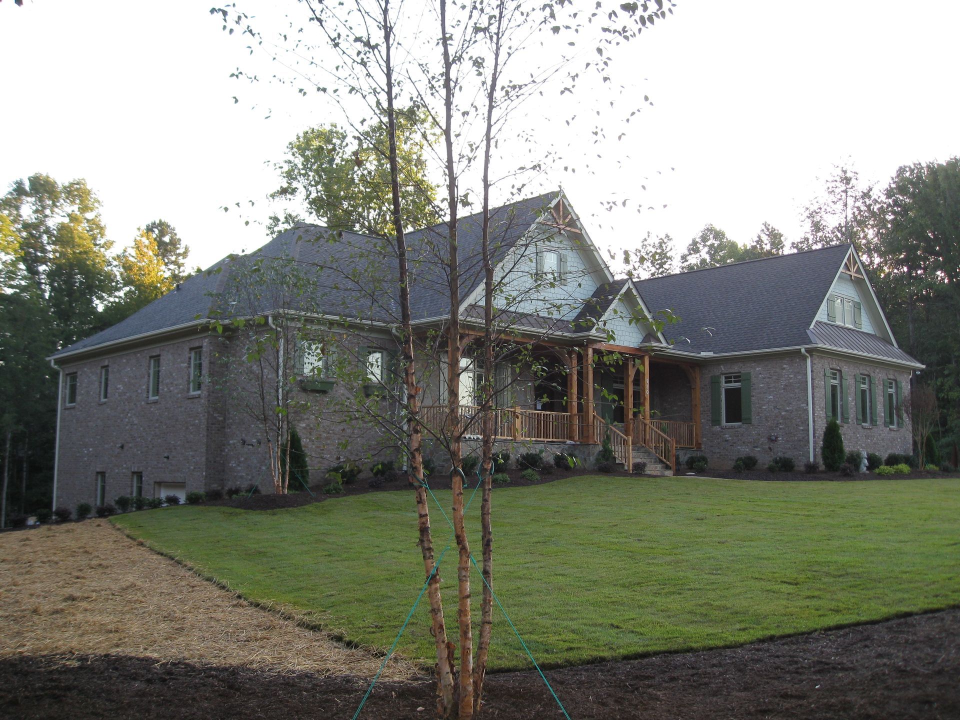 Stone house with dark roof and front porch on a grassy lawn, surrounded by trees.