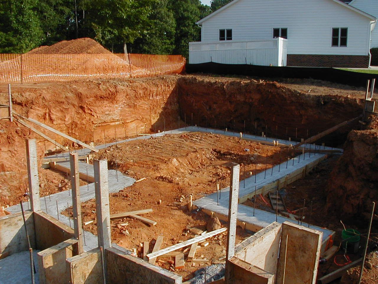 Excavated construction site with concrete foundation walls and a white house in the background