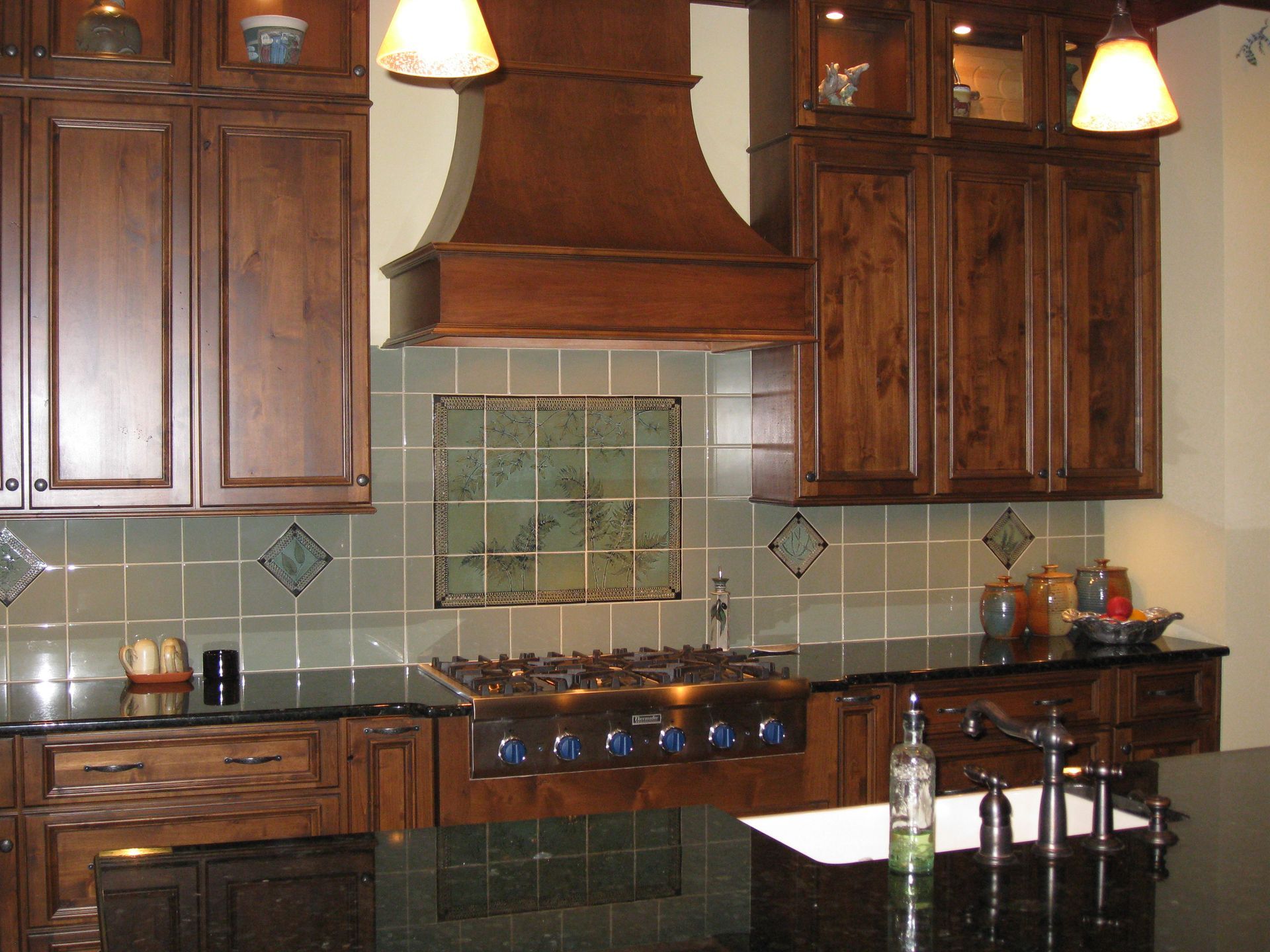 Warm kitchen with dark wood cabinets, tiled backsplash, stove, range hood, and hanging pendant lights