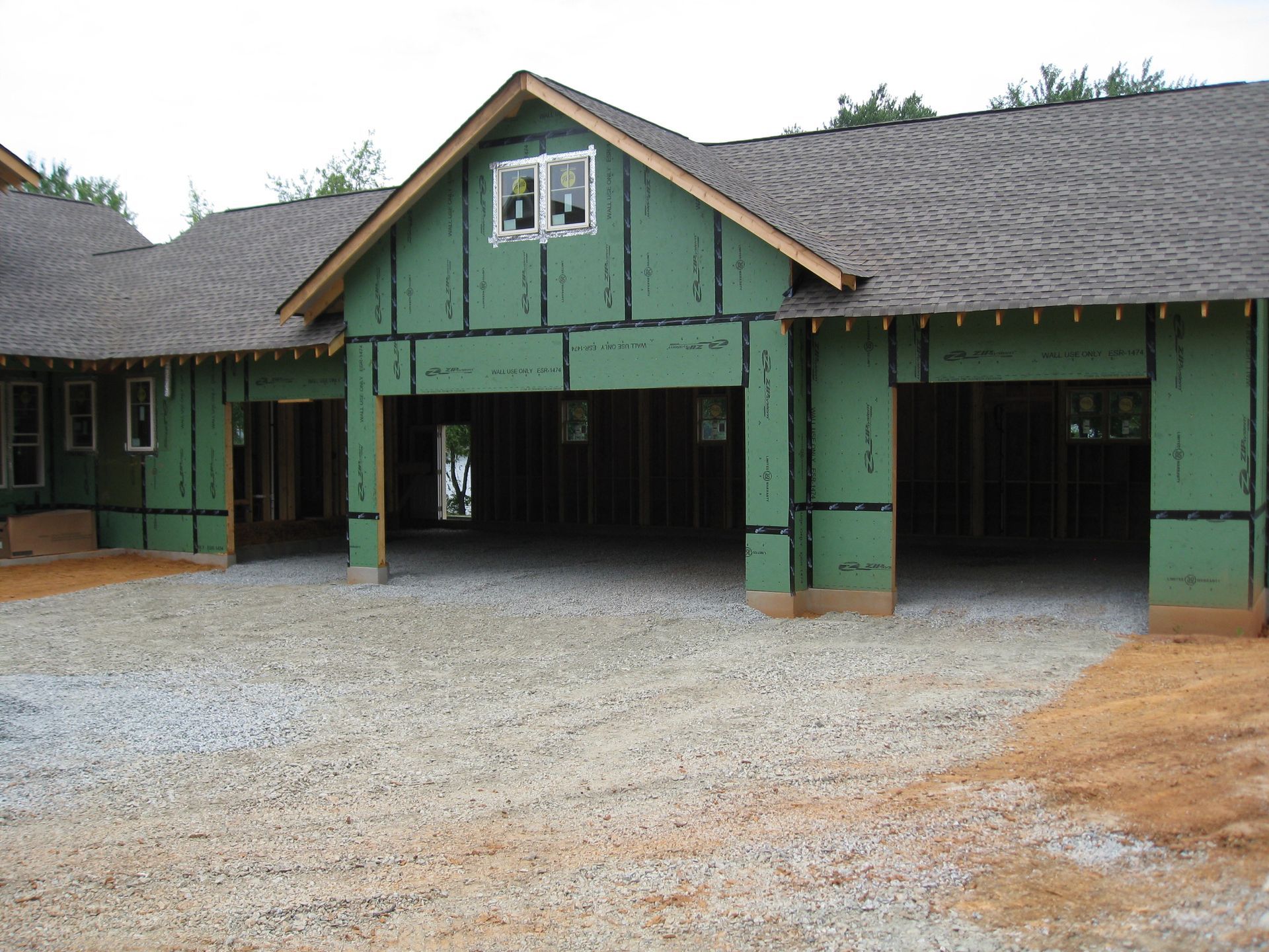 Green house under construction with three garage openings and a gravel driveway