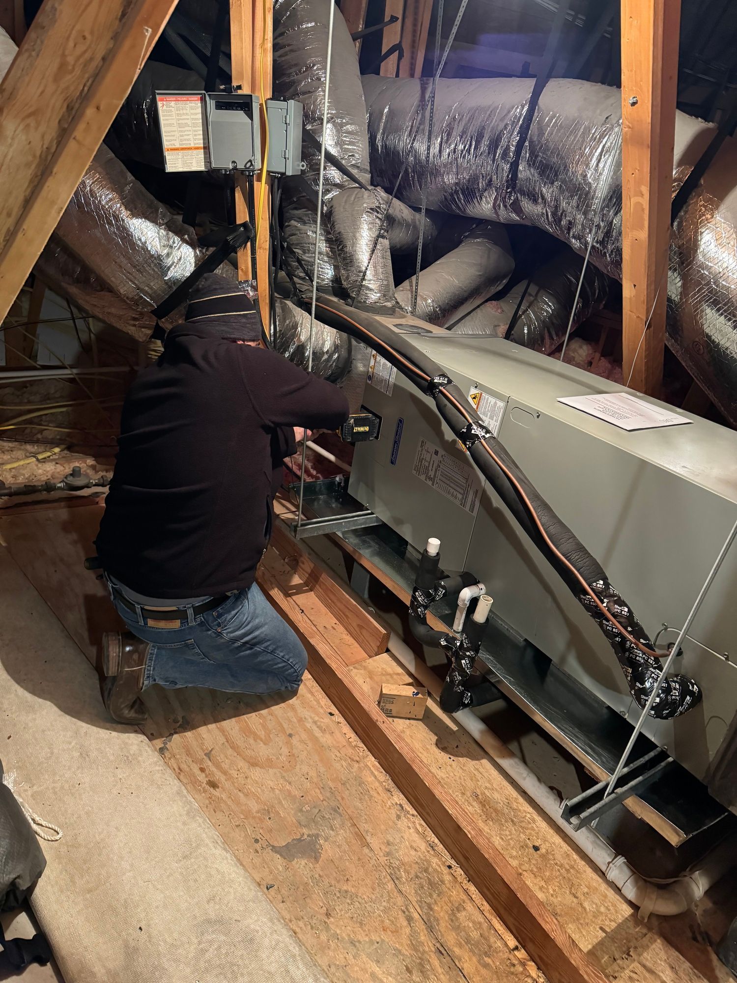 A person kneeling, working on an HVAC unit in an attic, surrounded by ductwork and wooden beams.