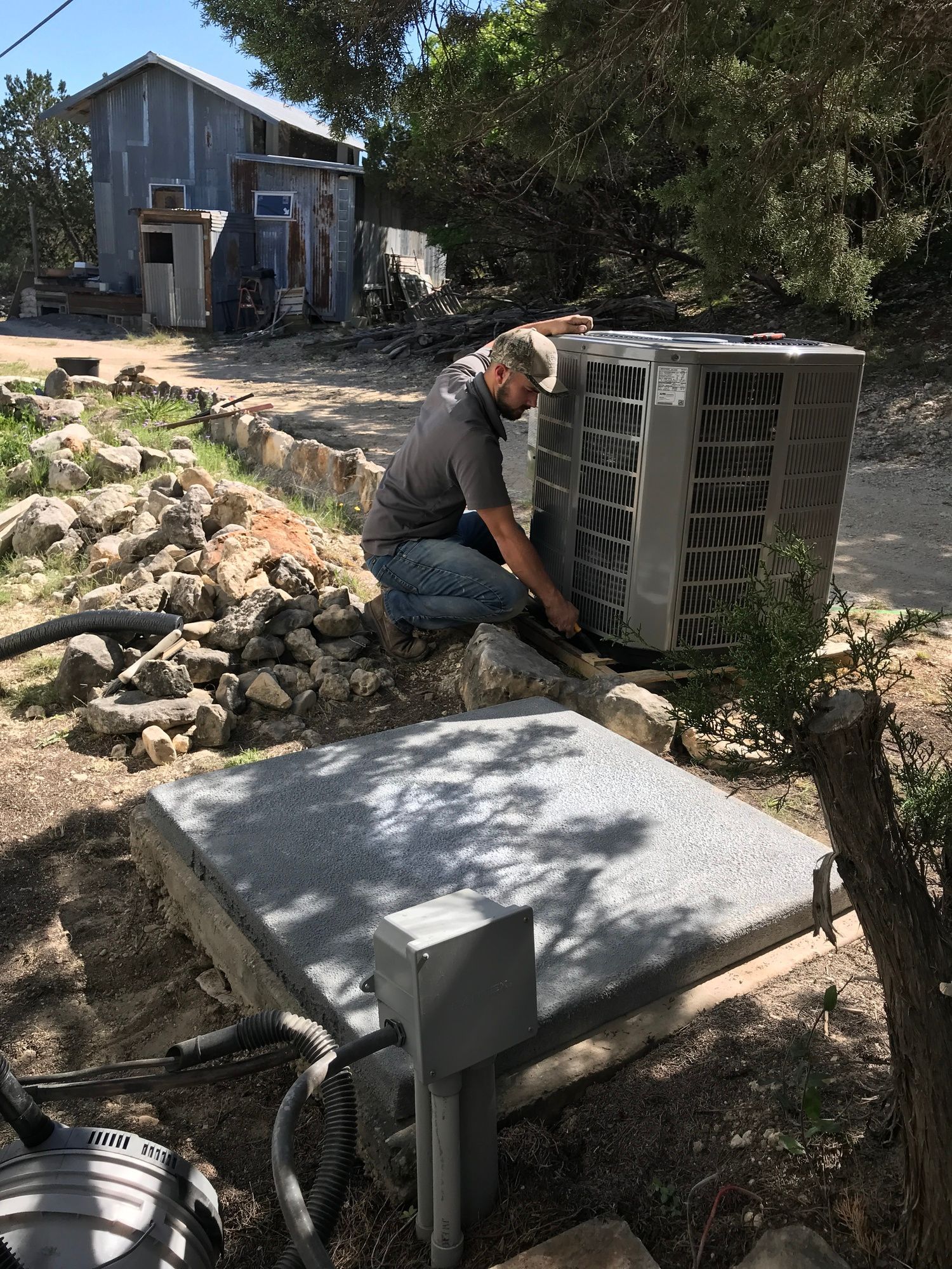 Man kneels near an air conditioner unit in a yard, prepping to install it. Building in the background.