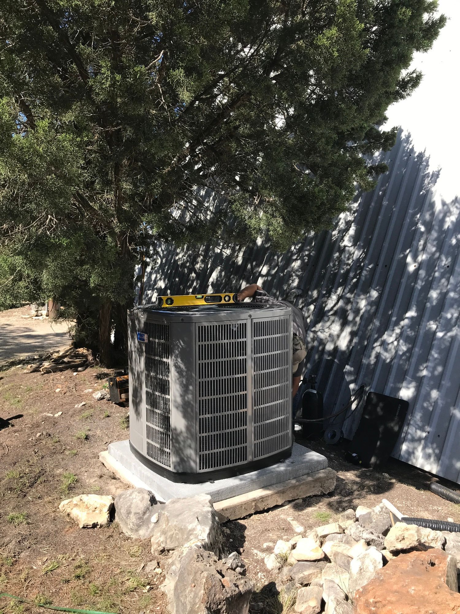 An air conditioning unit on a concrete base, near a tree and a metal shed.